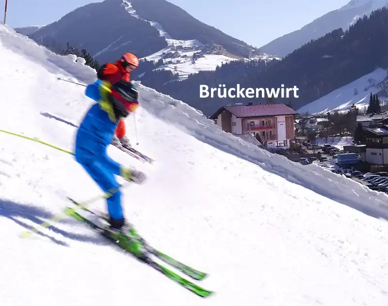 2 Schifahrer fahren auf der Schipiste direkt vor dem Haus Brueckenwirt